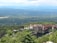 An expansive view of a scenic landscape featuring a large, historic resort complex nestled among lush greenery, with mountains in the background and a serene body of water in the foreground.