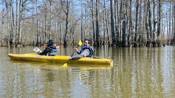 A yellow kayak glides along a tree-lined river, paddled by two people. One is actively rowing while the other is seated, looking away. Leafless trees rise from the water, casting reflections on the river's surface under a clear blue sky.
