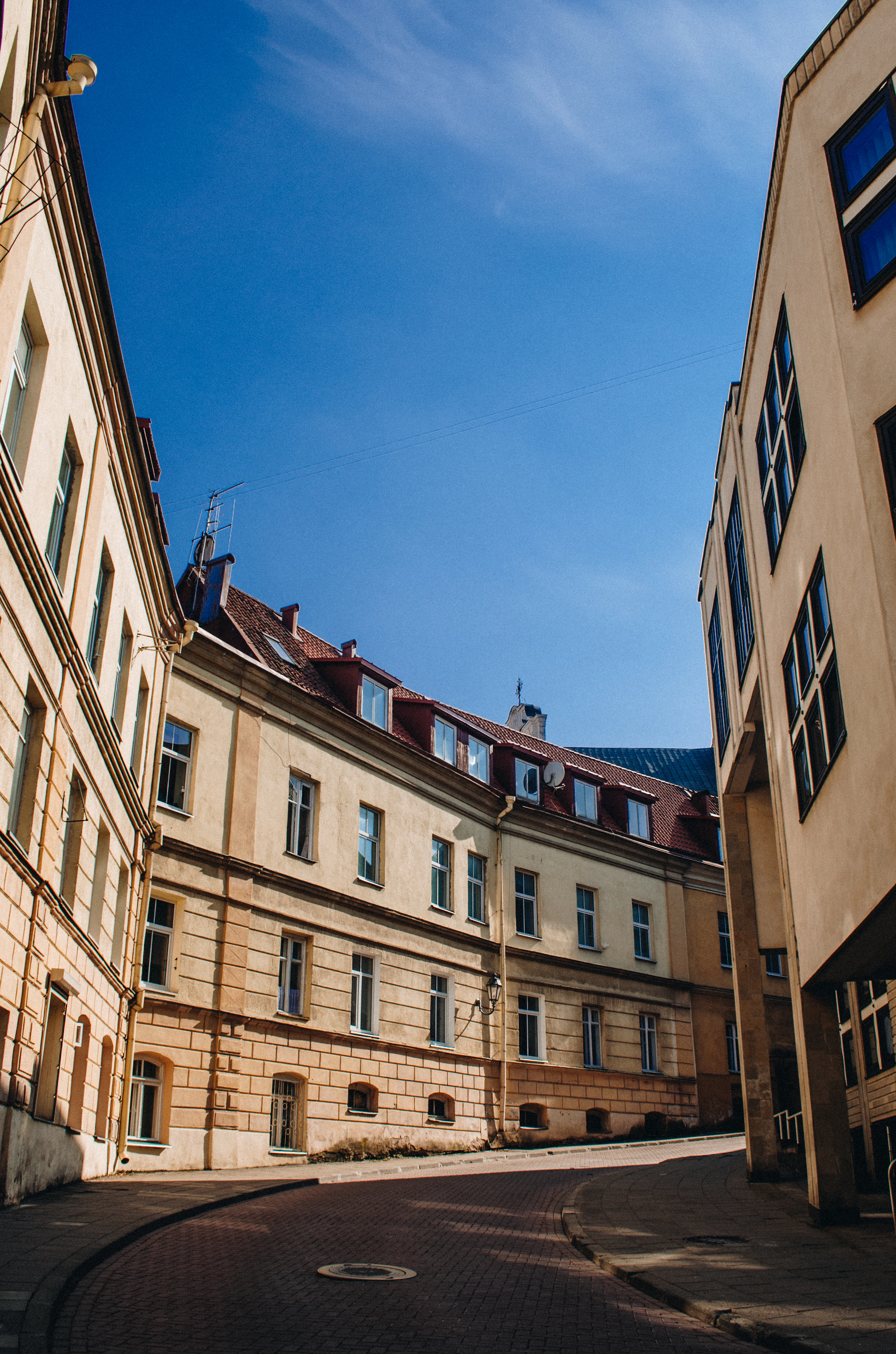 A curving cobbled street in Vilnius Old Town, warm afternoon sun raking across cream stucco facades and a red-tiled roof beneath a deep blue sky