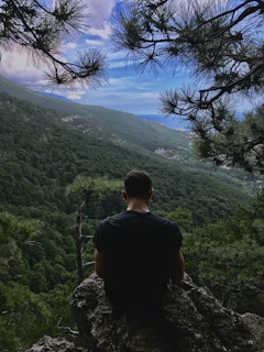 Iriel Thaleon assis en face d'une montagne et la mer entrain de réfléchir de dos