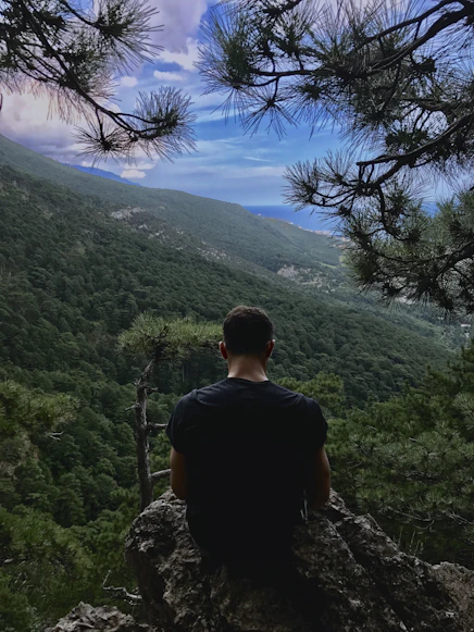 Iriel Thaleon assis en face d'une montagne et la mer entrain de réfléchir de dos