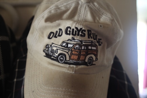 A beige cap with an embroidered design on the front featuring a vintage-style car and the text 'Old Guys Rule'. The cap appears slightly worn and is resting against a dark, patterned fabric.