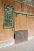 An old, weathered wall with a single metal-framed window featuring small glass panes. Next to the window, there's a sliding wooden door with graffiti. The setting appears to be an abandoned or neglected building with some wear and discoloration on the surfaces.