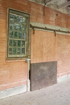 An old, weathered wall with a single metal-framed window featuring small glass panes. Next to the window, there's a sliding wooden door with graffiti. The setting appears to be an abandoned or neglected building with some wear and discoloration on the surfaces.