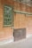 An old, weathered wall with a single metal-framed window featuring small glass panes. Next to the window, there's a sliding wooden door with graffiti. The setting appears to be an abandoned or neglected building with some wear and discoloration on the surfaces.