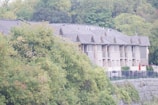 A row of modern townhouses with pitched roofs and large windows is partially obscured by lush green trees. The buildings are constructed with stone and wood materials and are surrounded by a fence. The setting appears to be a suburban area with dense greenery in the background.