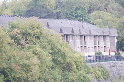 A row of modern townhouses with pitched roofs and large windows is partially obscured by lush green trees. The buildings are constructed with stone and wood materials and are surrounded by a fence. The setting appears to be a suburban area with dense greenery in the background.