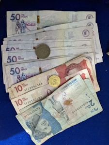 A collection of Colombian banknotes and coins is displayed on a dark blue surface. The notes are of different denominations, including 50,000, 10,000, and 2,000 pesos. Each note features distinct artwork and portraits. Two coins are visible, one silver and one gold in color.