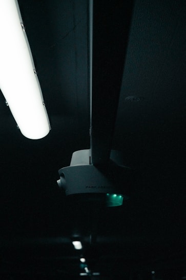A darkly lit scene featuring a ceiling-mounted light fixture and a sensor device. The light fixture emits a soft white glow, while the sensor has a small green light. The background is mostly in shadow, adding a sense of depth and mystery.