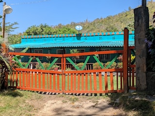 A colorful house is surrounded by lush greenery and hills. The building has a bright turquoise roof and walls with decorative patterns. A wooden gate painted in orange with green accents stands in the foreground. Potted plants with vibrant flowers line the entrance, and a satellite dish is mounted on top of the roof.