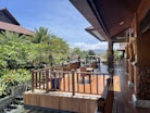 A spacious wooden terrace with intricate carvings overlooks a lush garden with palm trees. The clear sky and mountains are visible in the background. A table and chairs are arranged on the terrace for outdoor seating.