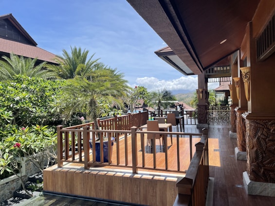 A spacious wooden terrace with intricate carvings overlooks a lush garden with palm trees. The clear sky and mountains are visible in the background. A table and chairs are arranged on the terrace for outdoor seating.