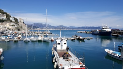 A picturesque marina with several boats docked at piers extending into calm blue waters. To the left, a quaint coastal town with pastel-colored buildings built on a hillside overlooks the harbor. The coastline and distant mountains provide a scenic backdrop under a clear blue sky.