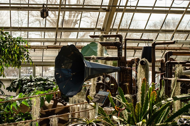 A greenhouse interior is filled with various plants and industrial equipment, including a large blue metal structure resembling a horn. Pipes and mechanical components are integrated with greenery, blending technology with nature. Sunlight filters through the glass ceiling, casting shadows over lush foliage and hanging mosses.
