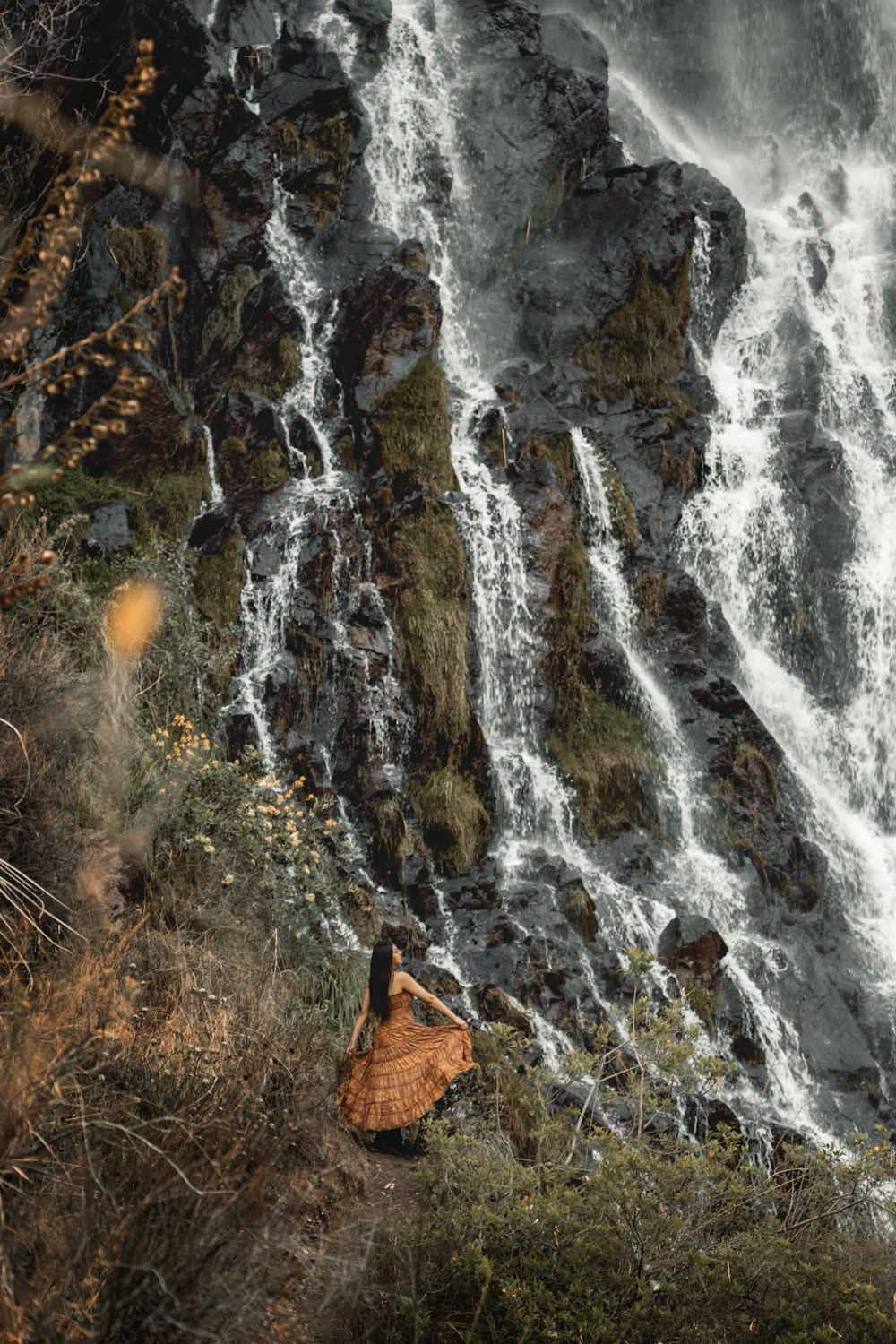 a woman in a brown dress standing in front of a waterfall