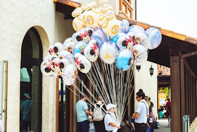 A vendor is holding a large bunch of colorful character-themed balloons featuring popular animated figures such as Minnie Mouse and characters from Toy Story. The scene is set against a backdrop of a building with stucco walls and arched windows. People are visible in the background, suggesting an outdoor shopping or amusement area.
