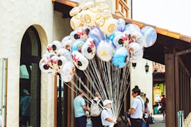 A vendor is holding a large bunch of colorful character-themed balloons featuring popular animated figures such as Minnie Mouse and characters from Toy Story. The scene is set against a backdrop of a building with stucco walls and arched windows. People are visible in the background, suggesting an outdoor shopping or amusement area.