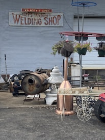 A welding shop with a variety of metal objects and tools is displayed. The ground is littered with industrial equipment, including a rusted metal drum, a decorative planter, and a red structure with hanging plants. The shop's sign is visible on the wall, suggesting the business specializes in welding and metalwork.