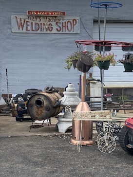 A welding shop with a variety of metal objects and tools is displayed. The ground is littered with industrial equipment, including a rusted metal drum, a decorative planter, and a red structure with hanging plants. The shop's sign is visible on the wall, suggesting the business specializes in welding and metalwork.