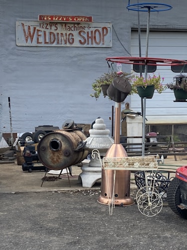 A welding shop with a variety of metal objects and tools is displayed. The ground is littered with industrial equipment, including a rusted metal drum, a decorative planter, and a red structure with hanging plants. The shop's sign is visible on the wall, suggesting the business specializes in welding and metalwork.