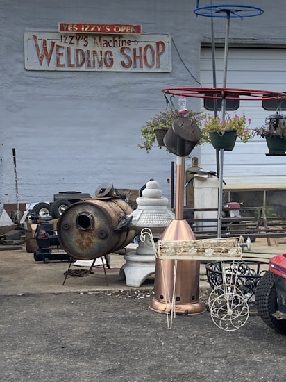 A welding shop with a variety of metal objects and tools is displayed. The ground is littered with industrial equipment, including a rusted metal drum, a decorative planter, and a red structure with hanging plants. The shop's sign is visible on the wall, suggesting the business specializes in welding and metalwork.