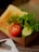 A sandwich served in a brown cardboard box rests on a wooden table. The sandwich includes a slice of toasted bread, fresh lettuce, a slice of ripe tomato, and a piece of cucumber. Next to the box, there is a metal fork placed on a folded white napkin.