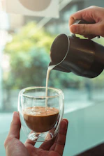Fresh milk being poured into a glass from a traditional metal jug on a wooden table surrounded by green fields.