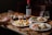 A wooden table set with an assortment of plates containing various meals, including pasta, quinoa, and vegetables, each topped with a poached egg. A wrapped sandwich sits on the table, and two glasses of white wine are present, one held by a hand. A bottle of rum with colorful labeling is placed centrally on the table. The background features a dark, leather-upholstered chair.