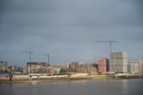 A riverside cityscape with modern construction projects visible. Several cranes stand tall amidst ongoing developments, including multiple buildings with a mix of architectural styles. The weather appears overcast, with a gray sky casting a muted light over the scene.