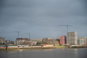 A riverside cityscape with modern construction projects visible. Several cranes stand tall amidst ongoing developments, including multiple buildings with a mix of architectural styles. The weather appears overcast, with a gray sky casting a muted light over the scene.
