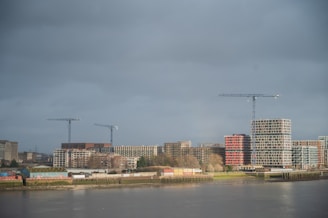 A riverside cityscape with modern construction projects visible. Several cranes stand tall amidst ongoing developments, including multiple buildings with a mix of architectural styles. The weather appears overcast, with a gray sky casting a muted light over the scene.