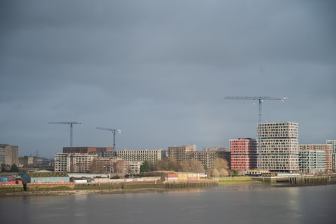 A riverside cityscape with modern construction projects visible. Several cranes stand tall amidst ongoing developments, including multiple buildings with a mix of architectural styles. The weather appears overcast, with a gray sky casting a muted light over the scene.