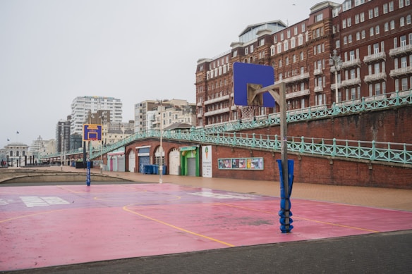 An outdoor basketball court with pink flooring and blue basketball hoops is situated next to a brick building with ornate architecture. The backdrop features multistory residential buildings and a seaside promenade with green railing and arches. The scene is overcast, giving it a calm and subdued feel.