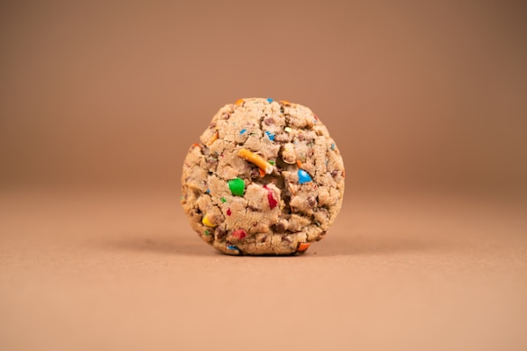 A single, round cookie with colorful candy pieces embedded throughout its crumbly texture, set against a smooth, light brown background.