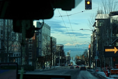 A street view from inside a vehicle, showing an urban scene with buildings on both sides, a few cars ahead, and snowy mountains in the distance under a partly cloudy sky. A traffic light is visible above, and an electronic arrow sign on the right.