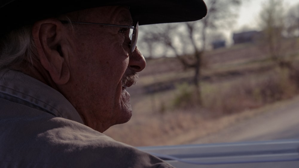 an older man wearing a hat and glasses driving a car