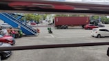 A parking area with several vehicles, including cars and a motorcycle. A large red shipping container is visible on the road, being transported by a truck. A blue staircase is seen on the left, leading to an elevated walkway. Trees line the far side of the street, and there are people near the parked vehicles.