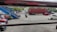 A parking area with several vehicles, including cars and a motorcycle. A large red shipping container is visible on the road, being transported by a truck. A blue staircase is seen on the left, leading to an elevated walkway. Trees line the far side of the street, and there are people near the parked vehicles.