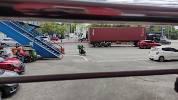 A parking area with several vehicles, including cars and a motorcycle. A large red shipping container is visible on the road, being transported by a truck. A blue staircase is seen on the left, leading to an elevated walkway. Trees line the far side of the street, and there are people near the parked vehicles.