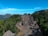 A traditional village with thatched-roof houses is nestled among lush green forests and mountains. The houses are arranged in two rows forming a path between them, and the area is surrounded by dense vegetation. The backdrop features a mountain range under a clear blue sky.