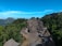 A traditional village with thatched-roof houses is nestled among lush green forests and mountains. The houses are arranged in two rows forming a path between them, and the area is surrounded by dense vegetation. The backdrop features a mountain range under a clear blue sky.