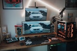 A modern gaming and computer setup featuring multiple monitors displaying an image of a car, a mechanical keyboard, gaming mouse, and a custom desktop PC with RGB lighting. The setup is on a wooden desk alongside professional audio equipment including speakers and a microphone mounted on a stand. Wall art and additional gadgets are visible in the background.