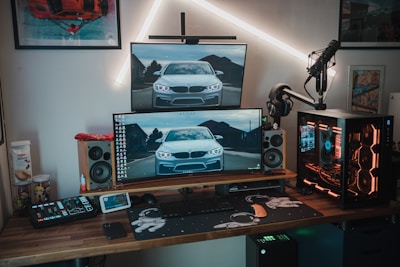 A modern gaming and computer setup featuring multiple monitors displaying an image of a car, a mechanical keyboard, gaming mouse, and a custom desktop PC with RGB lighting. The setup is on a wooden desk alongside professional audio equipment including speakers and a microphone mounted on a stand. Wall art and additional gadgets are visible in the background.