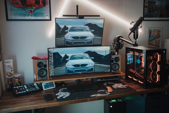 A modern gaming and computer setup featuring multiple monitors displaying an image of a car, a mechanical keyboard, gaming mouse, and a custom desktop PC with RGB lighting. The setup is on a wooden desk alongside professional audio equipment including speakers and a microphone mounted on a stand. Wall art and additional gadgets are visible in the background.