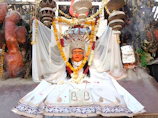 A vibrant religious statue adorned with a colorful floral garland, clothed in a white ceremonial dress. The backdrop features a variety of decorative elements, including hanging objects and intricate patterns. The scene is rich with cultural symbolism and detailed craftsmanship, evident in the paintings and embellishments.
