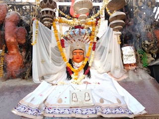 A vibrant religious statue adorned with a colorful floral garland, clothed in a white ceremonial dress. The backdrop features a variety of decorative elements, including hanging objects and intricate patterns. The scene is rich with cultural symbolism and detailed craftsmanship, evident in the paintings and embellishments.