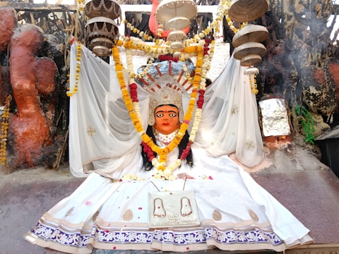 A vibrant religious statue adorned with a colorful floral garland, clothed in a white ceremonial dress. The backdrop features a variety of decorative elements, including hanging objects and intricate patterns. The scene is rich with cultural symbolism and detailed craftsmanship, evident in the paintings and embellishments.