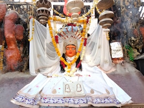 A vibrant religious statue adorned with a colorful floral garland, clothed in a white ceremonial dress. The backdrop features a variety of decorative elements, including hanging objects and intricate patterns. The scene is rich with cultural symbolism and detailed craftsmanship, evident in the paintings and embellishments.