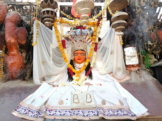 A vibrant religious statue adorned with a colorful floral garland, clothed in a white ceremonial dress. The backdrop features a variety of decorative elements, including hanging objects and intricate patterns. The scene is rich with cultural symbolism and detailed craftsmanship, evident in the paintings and embellishments.