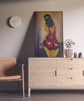 A modern interior space features a wooden chair and a sideboard. On the wall, there is a circular white clock with minimalistic numbers. A large painting leans against the wall, depicting a woman in a vibrant red dress seen from behind, holding a small object. The sideboard holds a decorative plant in a copper pot and two black bookends.